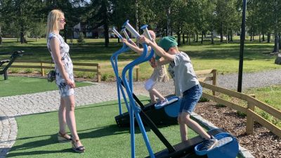 En kvinna med sommarklänning står med en liten flicka och en liten pojke som testar gymredskap