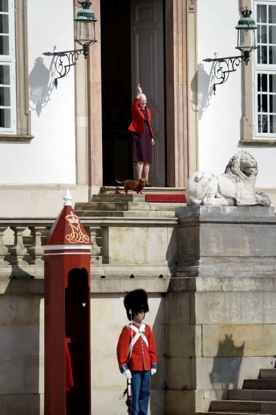 Drottning Margrethe II vinkar på trappan till ett slott i sällskap av en tax. Nedanför trappan står en vakt i röd rock och bäverskinnsmössa.