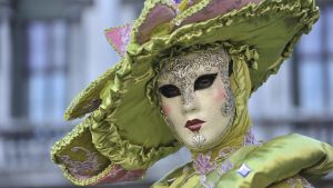 Vihreän ja roosan sävyiseen naamiaisasuun ja päähineeseen sekä taidokkaasti koristeltuun naamioon pukeutunut henkilö Venetsian karnevaaleissa.