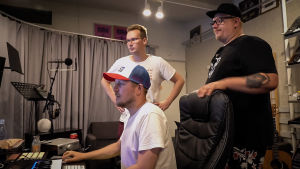 Unelmia ja studiohommia. Kolme miestä musiikkistudiossa. Yksi istuu äänipöydän edessä, kaksi seisoo hänen takanaan.