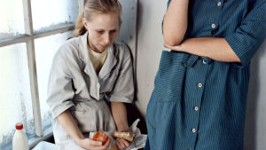 Iris (Kati Outinen) har det inte lätt. Iris (Kati Outinen) sitter på ett fönsterbräde med en smörgås i handen och ser uppgiven ut.