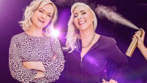 Blondit! Kaksi blondia katsoo ja hymyilee kameraan violettia taustaa vasten. Toisen hiuksiin suihkutetaan sivusta hiuslakkaa.