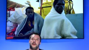 Italiens inrikesminister Matteo Salvini förde en hård migrationspolitik Italiens inrikesminister Matteo Salvini framför en bild av Medelhavsflyktingar.
