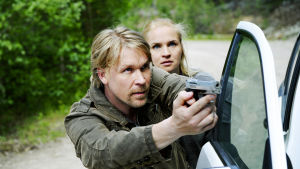 Mies ja nainen kyyryssä auton oven vieressä. Mies osoittaa aseella pois kuvasta, nainen hänen takanaan. Sarjasta Pirunpelto.