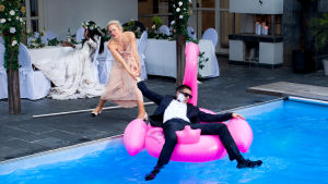 Man i kostym ligger avtuppad på en uppblåsbar flamingo i en pool. Han fru försöker dra upp honom.