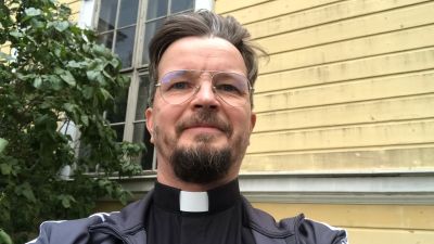 Papin omakuva puun ja keltaisen rakennuksen edessä