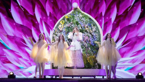 Greklands Katerine Duska i Eurovisionen 2019. Greklands Katerine Duska framför en blomstervägg med tre vitklädda dansare.