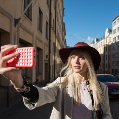 Donna (Alina Tomnikov) står på gatan och håller en telefon med framsträckt hand framför ansiktet. Solen skiner i ansiktet på Donna och i bakgrunden syns bilar.