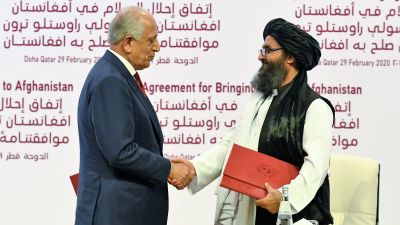 Khalilzad ja Baradar kättelevät. Khalilzadilla on tummansininen puku ja viininpunainen kravatti. Baradarilla on musta turbaani ja perinteinen asu. Hänellä on hieman harmaantunut parta ja silmälasit.