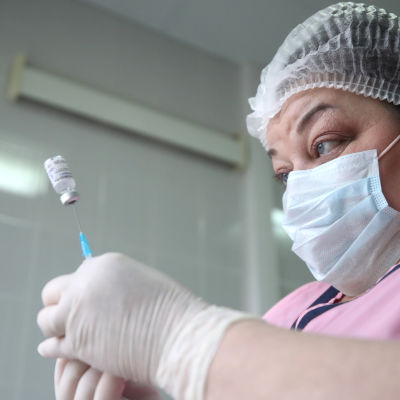 Sjukskötare med ansiktsmask sticker en nål in i en liten glasburk med vaccin.