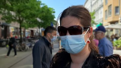 Katja Hedman, en dam med långt mörkt hår i hästsvans, solglasögon och ljusblått munskydd, står på en gågata en solig sommardag.