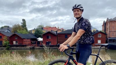 En man står med en cykel och tittar leende in i kameran. Bakom honom syns Borgå å och röda träbodar.