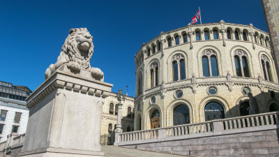 Leijonapatsas jalustalla parlamenttitalon edessä. Norjan lippu liehuu talon katolla.