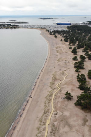 Från luften kan man se hur stråket ringlar sig fram bland dynerna. Ett flygfoto över en strand där man ser havet på ena sidan och skogen på andra. I sanden har ett promenadstråk i trä byggts. Uppe i bild syns en hamn med ett fartyg.