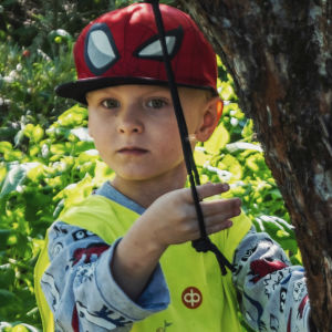 Punaiseen lippalakkiin ja keltaiseen huomioliiviin pukeutunut pikkupoika seisoo keskellä vehreää puutarhaa ja leikkii narulla, joka roikkuu omenapuusta.