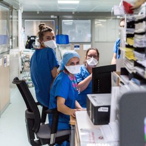 Sjukskötare i munskydd tittar på en datorskärm i ett arbetsrum med en massa papper. 