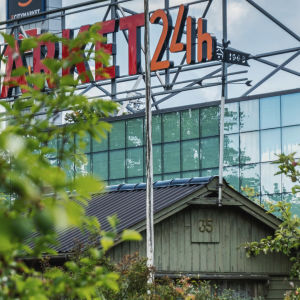 Edustalla omenapuita ja vanha, vihreä mökki. Taustalla suuri lasiseinä ja kyltti, jossa punaisella teksti "Citymarket 24h".