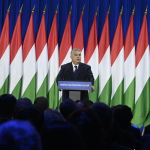 Viktor Orbán står vid ett talarpodium framför en lång rad ungerska flaggor. I förgrunden skymtar huvuden i publiken.