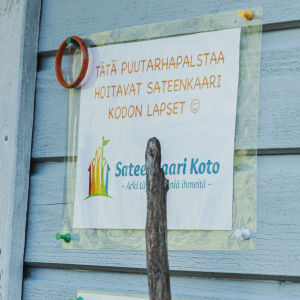 Vaaleansininen talonseinä, kyltissä teksti, jossa lukee että "tätä puutarhapalstaa hoitavat Sateenkaari Kodon lapset". 