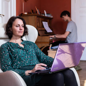 En kvinna sitter i en fåtölj och jobbar. Hon har en bärbar dator i famnen. I bakgrunden syns en yngre man som spelar piano.