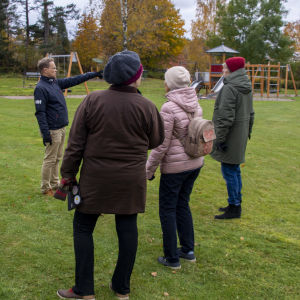 Människor står i en grupp på en gräsmatta. En man i mitten står och pekar. I bakgrunden syns en lekplats.