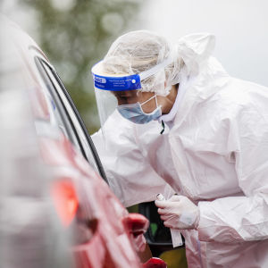 En person utför ett coronatest på en patient som sitter i sin bil (endast bilen syns på bilden). Persoenn som utför testet har skyddsdräkt på sig.