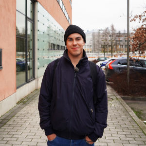En ung man i jacka och mössa ler in i kameran framför parkerade bilar och en husvägg.