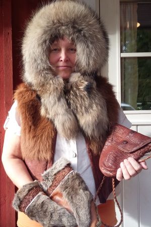 Nina Karlsson iklädd en mårdhundsmössa, kalvskinnsväst och i handen håller hon en väska gjord av pungen av ett djur. Nina Karlsson iklädd sina skinnprodukter.
