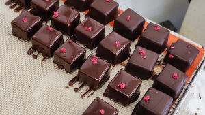 De här chokladkonfekterna har just fått sin skinande yta. Fyrkantiga chokladkonfekter som just doppats i smält choklad. Ovanpå dem ligger små röda blomblad.