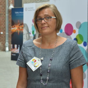 Eva Österbacka, professor i ekonomi vid Åbo Akademi och ordförande för konferensens organisationskommitté. Professor Eva Österbacka i en grå klänning framför planscher och en teglad vägg.