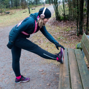 Liisa-Maija Huttunen i träningskläder och reflexsele. Hon står och stretchar benet mot en bänk i en skog.