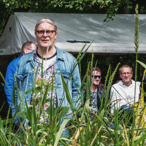 Yleisöä kuuntelemassa ulkoilmakonsertissa, taustalla valkoinen telttakatos, etualalla korkeaa ruohikkoa.