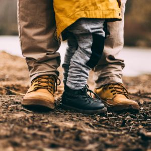 En pappas och ett barns fötter och skor. Barnet står mellan pappans ben.