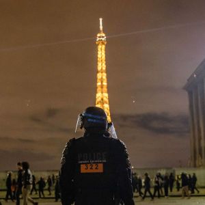 Det är mörkt. En polis står med ryggen mot kameran och skymmer delvis Eiffeltornet i bakgrunden. Människor promenerar förbi..  
