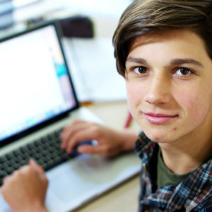 Kuvassa on tyytyväisen näköinen nuori poika tietokoneen äärellä. 
