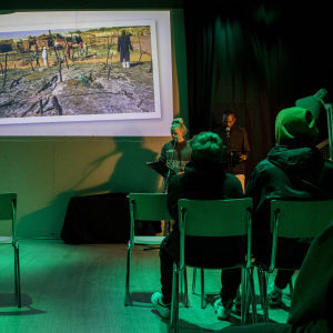 I ett mörkt rum visas ett fotografi från ett flyktingläger. I publiken sitter åttondeklassare.