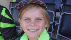 Ember gillar Forest Green Rovers. En ung och glad flicka sitter på en fotbollsläktare. Hon har en ljusgrön halsduk runt sin hals.