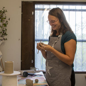En kvinna med förkläde på står i en keramikverkstad. Hon håller på och formar en klump med lera som hon håller i händerna.
