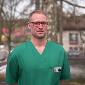 Man i 40-årsåldern med kort hår, glasögon, grön sjukskötarblus och vita byxor.