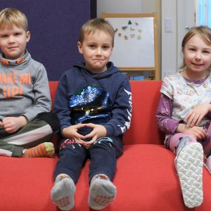 Bilden föreställer tre glada förskolebarn som sitter i en röd soffa.