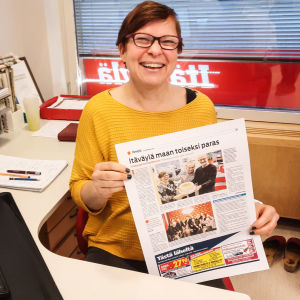 En kvinna i gul tröja sitter vid ett skrivbord. Hon håller upp en tidningssida mot kameran och ler.