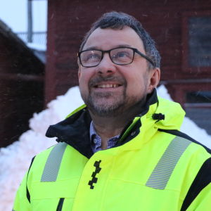 En man med glasögon och neonjacka blickar uppåt mot skyn. Bakom honom skymtar en stor snöhög och röda träbyggnader.
