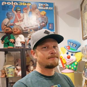 En man med keps och blond skäggväxt står framför en vägg fylld med spel och leksaker. På väggen finns ett bordshockeyspel tillverkat av företaget Bock-Plast.