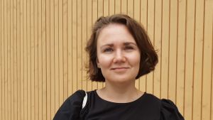 Roosa Veijola hade det svårt med distansjobbet i början. Ung kvinna i brunt hår till hakan, svart skjorta, står framför en brun trävägg.