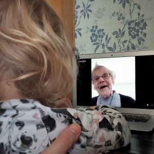 Ett barn med lockigt hår ser på en dataskärm med en bild på en äldre kvinna som ler. 