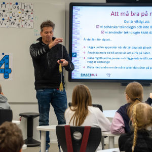 En man står framför en klass med skolelever. Han håller i två pinnar med tofsar och håller på med att visa ett trolleritrick.