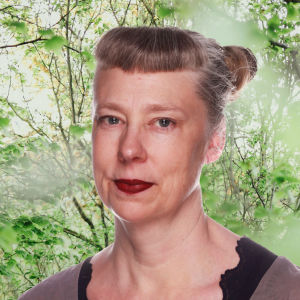 En ljushårig kvinna ser rakt in i kameran. Håret är uppsatt i en tofs och i bakgrunden syns en grön trädgård.