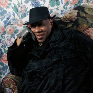 Muotitoimittaja André Leon Talley istuu mustassa viitassa kukikkaan tapetin peittämän seinän edessä. Kuva dokumenttielokuvasta.
