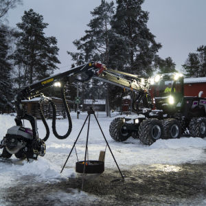 En Logset skogsprocessor står på en snöig gårdsplan med lamporna tända. 
