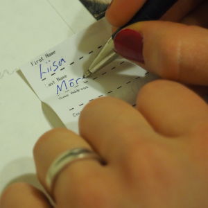 Nainen kirjoittaa nimeään paperiin, häiden kiitoskortti taustalla.